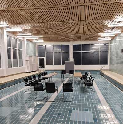 Ein leeres Schwimmbecken in einer Halle. Auf dem Boden des Beckens stehen Stühle.